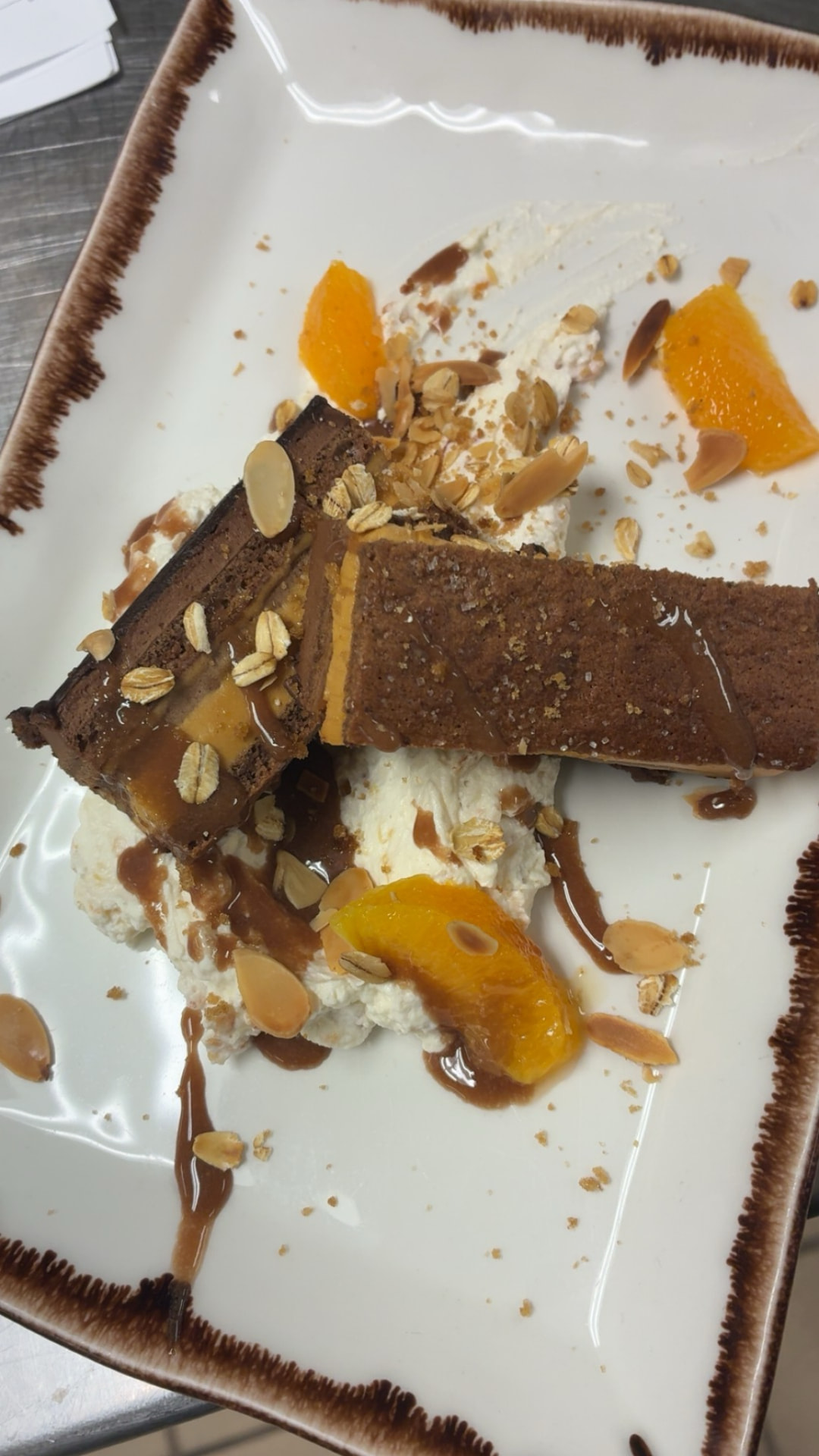 Chocolate dessert slices with orange segments, cream, sliced almonds, oats and brown sauce on a rectangular plate