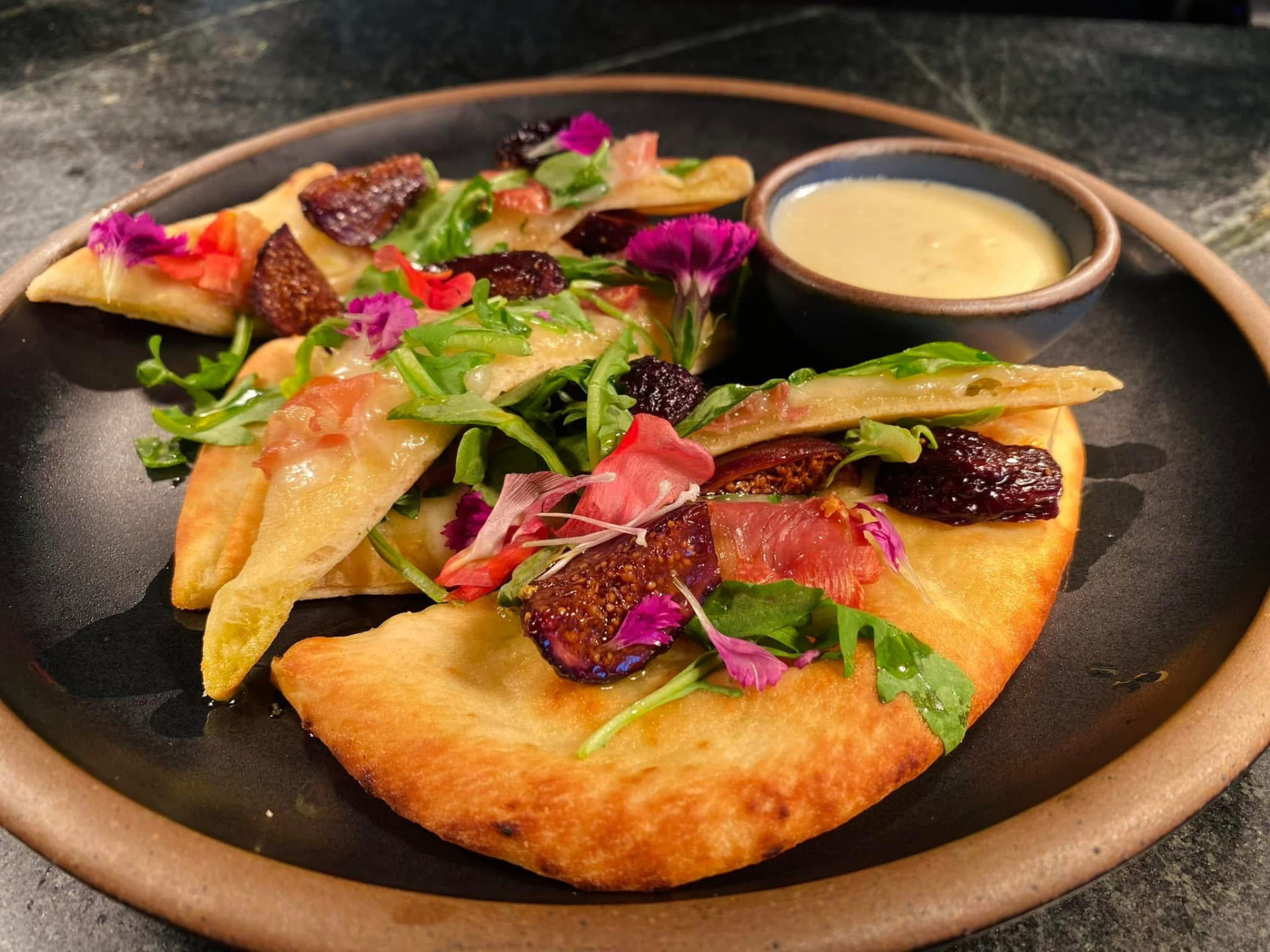 Golden flatbread wedges topped with figs, cured meat, greens and colorful flowers beside a bowl of pale sauce